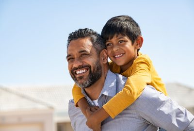 Happy middle eastern child enjoying ride on father back outdoor. Smiling young dad giving piggyback ride to son on street while looking away with copy space. Latin cheerful man carrying on shoulder indian kid: future and vision concept .
